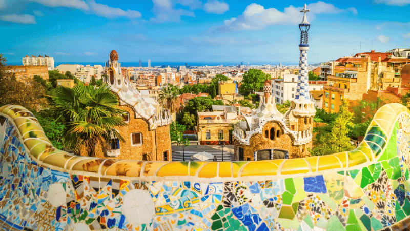 Barcelona skyline from Park Güell with Gaudí’s mosaic terrace and historic architecture under a blue Mediterranean sky.