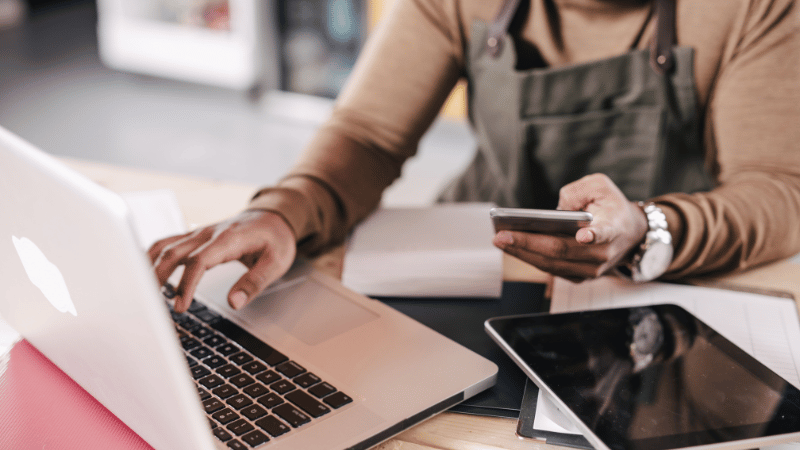 A small business owner wearing an apron works on a laptop while holding a smartphone, representing the process of managing and refinancing business loans online.