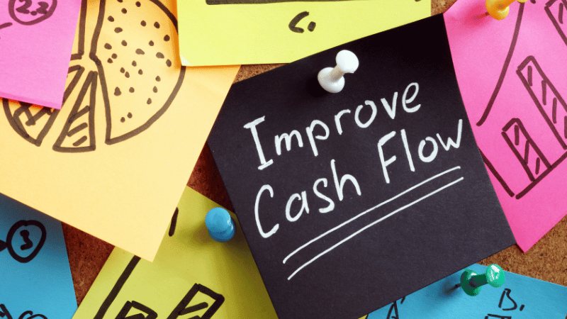 A black sticky note with 'Improve Cash Flow' pinned to a corkboard with other notes showing charts, representing the goal of using a business loan for better cash flow management.