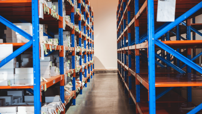 An aisle in a warehouse with tall blue and orange shelves, one side stocked with white boxes and the other side empty, representing inventory management.