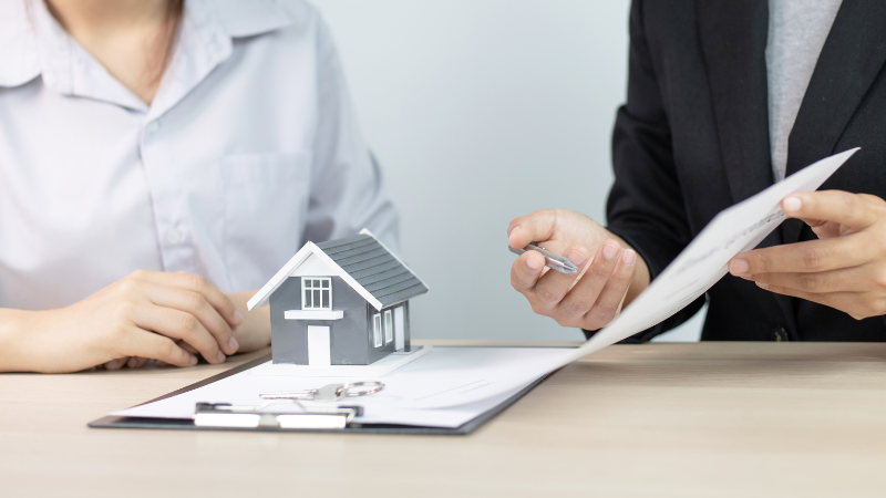 A professional mortgage advisor explains the terms of a fixed or variable rate mortgage document to a client, with a model house and keys on the table.