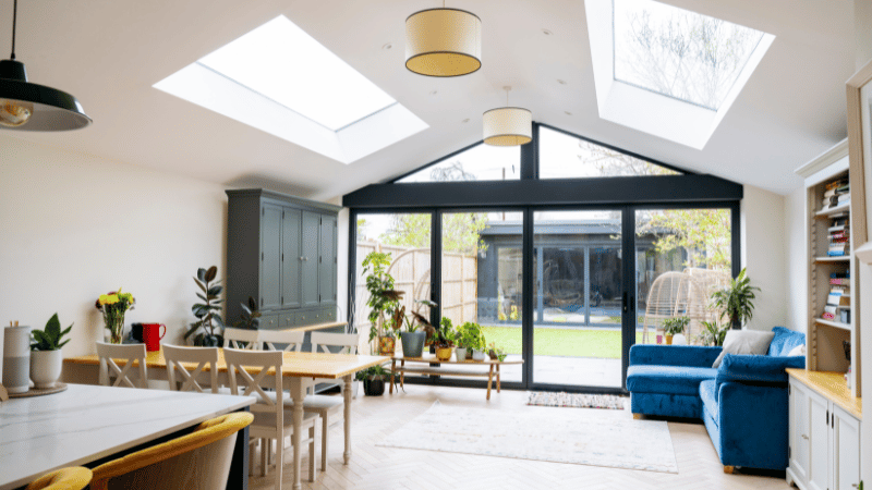 Bright and modern open-plan house extension with a vaulted ceiling, skylights, and large glass doors leading to a garden. The space includes a dining area and a living area with a blue sofa.