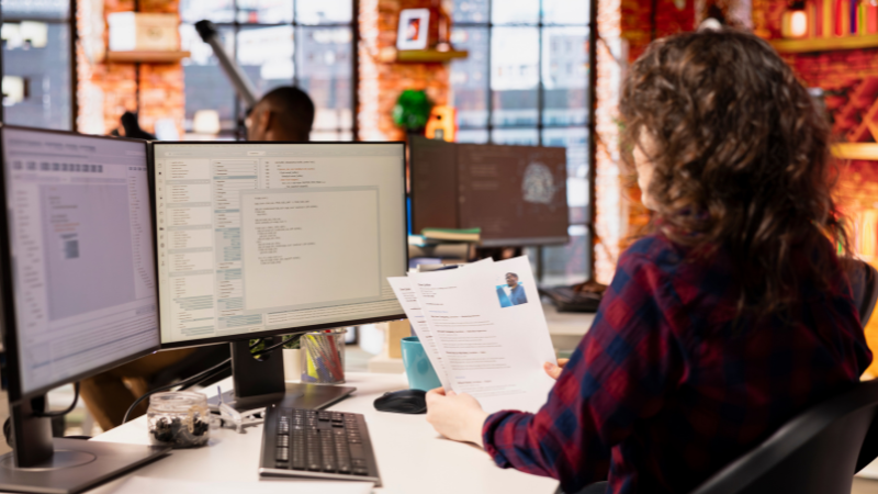 A recruitment professional at a modern office desk, reviewing a candidate's CV with several computer screens showing code in the background.