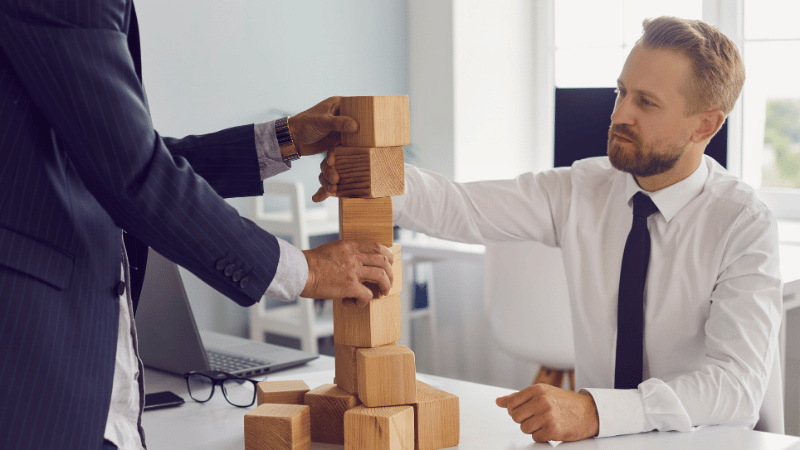 Two business professionals building a wooden block tower representing the steps, timelines, and funding layers of the UK management buyout process