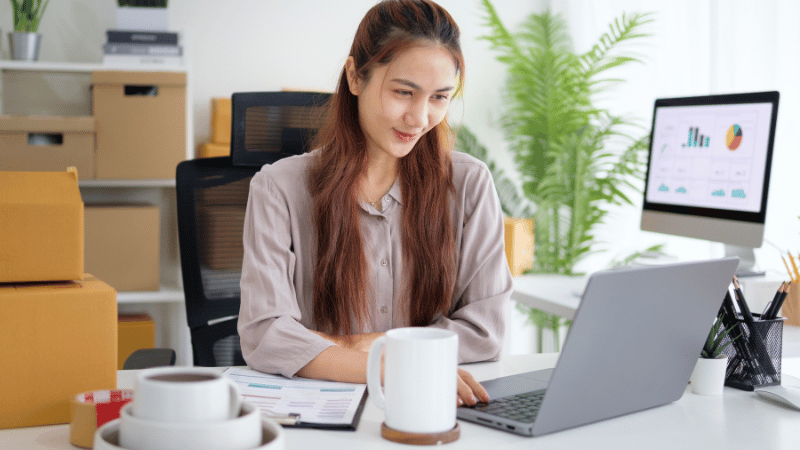A female eCommerce business owner smiling while working on a laptop at her desk, surrounded by shipping boxes and with business charts on a monitor behind her.