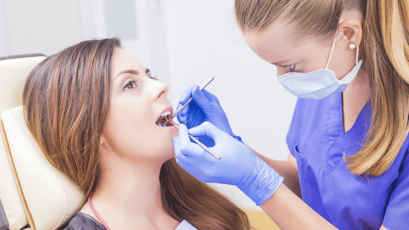 A female dentist wearing a mask and blue scrubs carefully performs a check-up on a female patient in a dental chair.