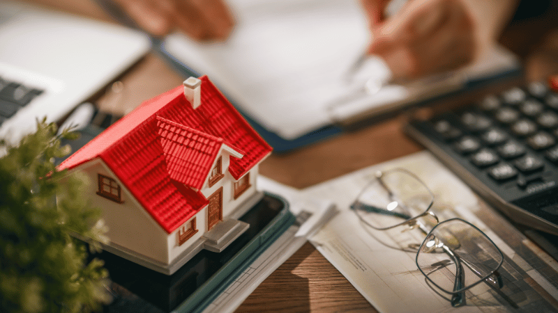 A close-up of a person signing a property development loan document, with a model house, calculator, and architectural plans on the desk.