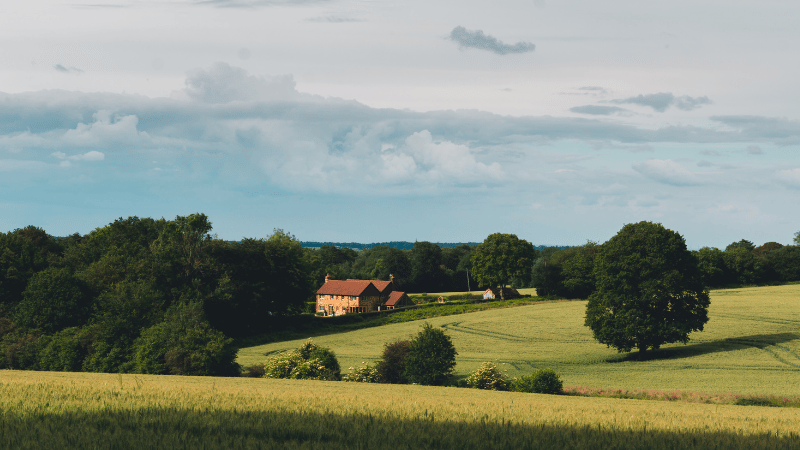 A traditional country house with a red roof nestled amongst trees in rolling green and golden fields under a wide, cloudy sky.