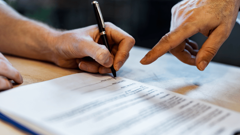 A close-up of a person signing a self-assessment tax loan document while a financial advisor points to the signature line.